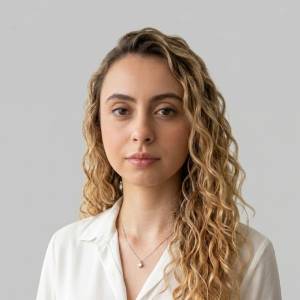 Uma pessoa jovem com cabelos longos e ondulados, usando uma blusa branca e um colar discreto. Ela está olhando levemente para o lado, com expressão neutra, diante de um fundo cinza claro.